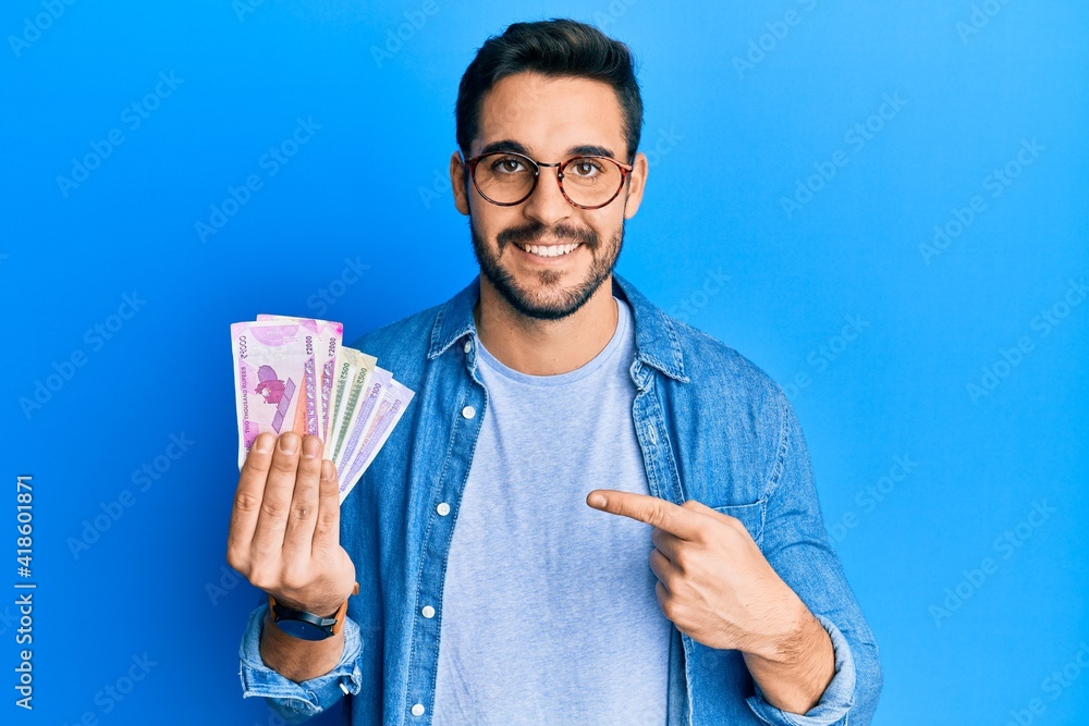 Wall mural young hispanic man holding indian rupee banknotes smiling happy pointing with hand and finger