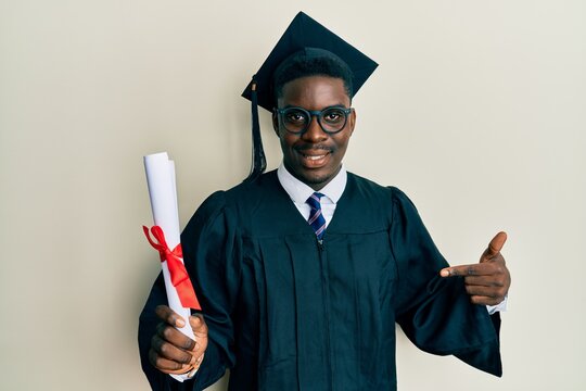Handsome black man wearing graduation cap and ceremony robe holding diploma pointing finger to one self smiling happy and proud