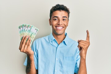 Young handsome african american man holding 50 hong kong dollars banknotes smiling with an idea or question pointing finger with happy face, number one