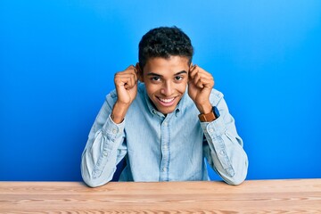 Young handsome african american man wearing casual clothes sitting on the table smiling pulling...