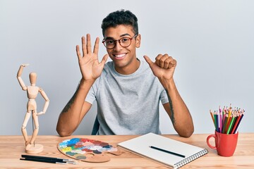 Young handsome african american man painter sitting palette and art manikin showing and pointing up with fingers number six while smiling confident and happy.
