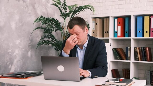 Eye fatigue, poor eyesight, office work. The young man takes off his glasses and rubs his eyes with his hand sitting at a table with a laptop in a suit. He closes his laptop, gets up and leaves the