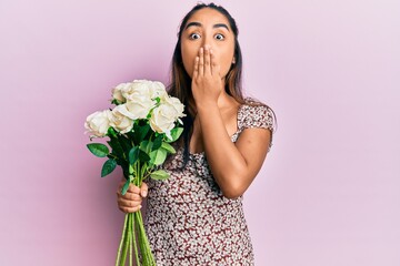 Young latin woman holding flowers covering mouth with hand, shocked and afraid for mistake....