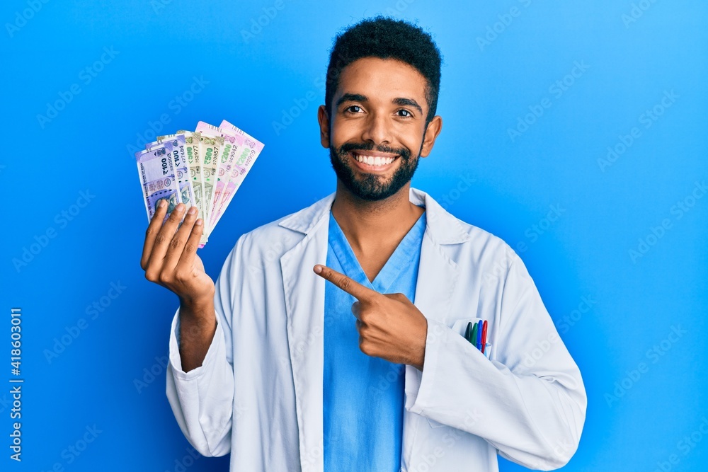 Canvas Prints handsome hispanic man with beard wearing medical uniform holding indian rupee smiling happy pointing