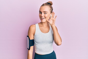 Beautiful blonde sport woman wearing arm band and earphones smiling with hand over ear listening and hearing to rumor or gossip. deafness concept.