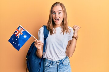 Beautiful blonde woman exchange student holding australia flag pointing thumb up to the side smiling happy with open mouth © Krakenimages.com