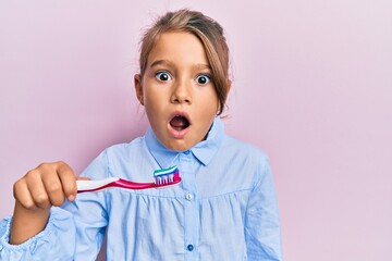 Little beautiful girl holding toothbrush with toothpaste scared and amazed with open mouth for...