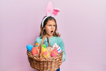 Little beautiful girl wearing cute easter bunny ears holding wicker basket with colored eggs angry and mad screaming frustrated and furious, shouting with anger. rage and aggressive concept.