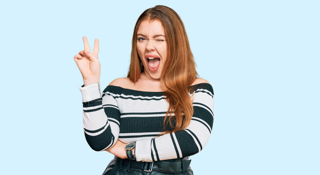 Young beautiful redhead woman wearing elegant clothes smiling with happy face winking at the camera doing victory sign. number two.