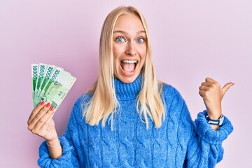 Young blonde girl holding 10000 south korean won banknotes pointing thumb up to the side smiling happy with open mouth