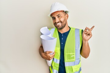 Handsome man with beard wearing safety helmet holding blueprints smiling happy pointing with hand and finger to the side