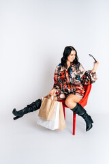 Happy smiling beautiful girl with shopping bags sitting on a red chair white background short dress mock up copy space hold package bag black overknees