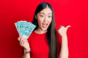 Young chinese woman holding 50 yuan chinese banknotes pointing thumb up to the side smiling happy with open mouth