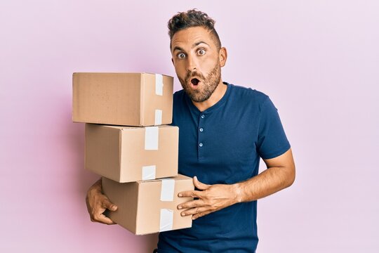 Handsome man with beard holding delivery packages scared and amazed with open mouth for surprise, disbelief face