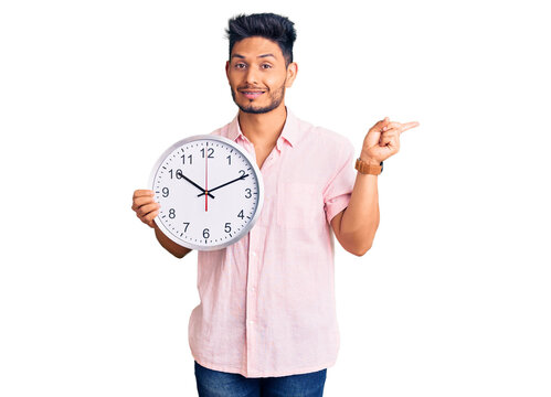 Handsome latin american young man holding big clock smiling happy pointing with hand and finger to the side