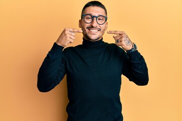 Handsome man with tattoos wearing turtleneck sweater and glasses smiling cheerful showing and pointing with fingers teeth and mouth. dental health concept.