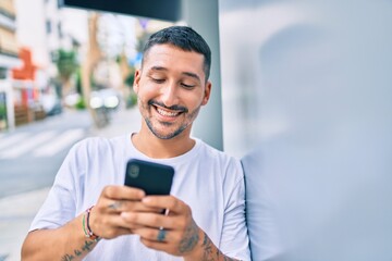 Young hispanic man smiling happy using smartphone leaning on the wall
