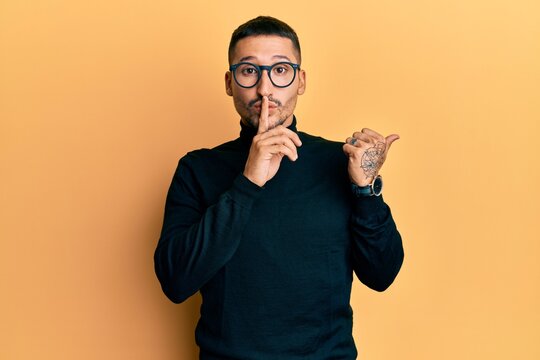 Handsome man with tattoos wearing turtleneck sweater and glasses asking to be quiet with finger on lips pointing with hand to the side. silence and secret concept.