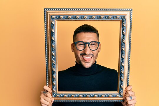 Handsome man with tattoos holding empty frame sticking tongue out happy with funny expression.