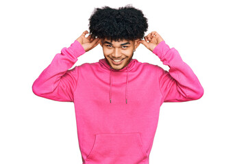 Young african american man with afro hair wearing casual pink sweatshirt smiling pulling ears with...