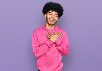 Obraz na płótnie Canvas Young african american man with afro hair wearing casual pink sweatshirt smiling with hands on chest with closed eyes and grateful gesture on face. health concept.