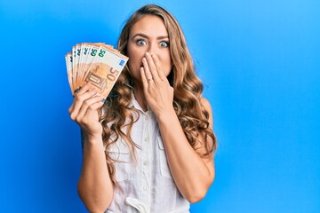 Young blonde girl holding euro banknotes covering mouth with hand, shocked and afraid for mistake....
