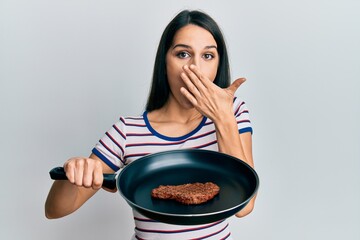 Young hispanic woman holding skillet with beef steak covering mouth with hand, shocked and afraid...