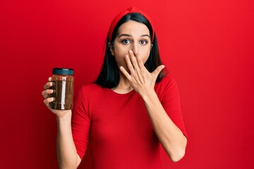 Young hispanic woman holding soluble coffee covering mouth with hand, shocked and afraid for...