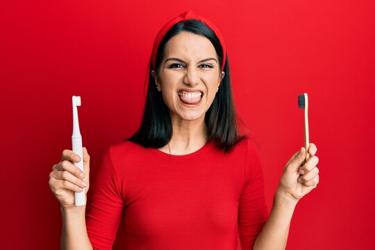 Young hispanic woman choosing electric toothbrush or normal teethbrush sticking tongue out happy with funny expression.