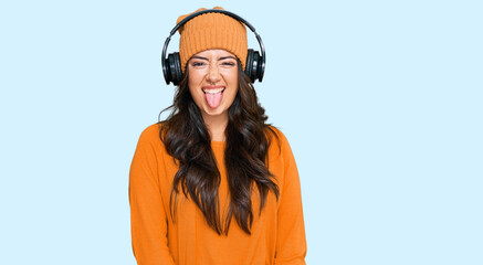 Beautiful brunette young woman listening to music using headphones sticking tongue out happy with funny expression. emotion concept.