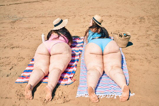Backview of two plus size overweight sisters twins women relaxing lying on a towel at the beach on summer holidays