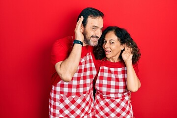 Middle age couple of hispanic woman and man wearing cook apron smiling with hand over ear listening an hearing to rumor or gossip. deafness concept.