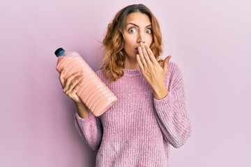 Young caucasian woman holding laundry softener bottle covering mouth with hand, shocked and afraid...