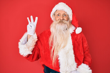 Old senior man with grey hair and long beard wearing traditional santa claus costume showing and pointing up with fingers number three while smiling confident and happy.