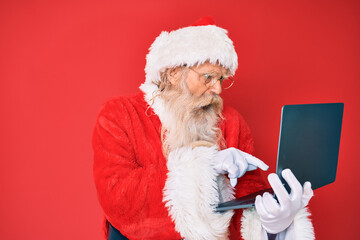 Old senior man with grey hair and long beard wearing santa claus costume using laptop angry and mad screaming frustrated and furious, shouting with anger. rage and aggressive concept.