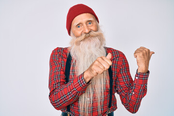 Old senior man with grey hair and long beard wearing hipster look with wool cap pointing to the back behind with hand and thumbs up, smiling confident