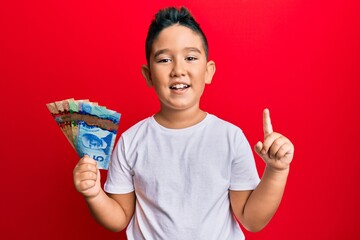 Little boy hispanic kid holding canadian dollars smiling with an idea or question pointing finger...