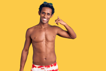 African handsome man wearing swimsuit and sunglasses smiling doing phone gesture with hand and fingers like talking on the telephone. communicating concepts.