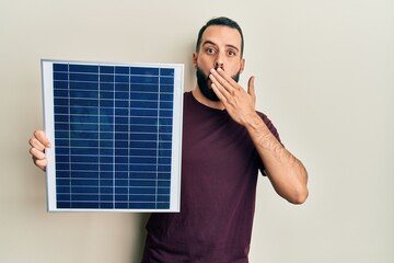 Young man with beard holding photovoltaic solar panel covering mouth with hand, shocked and afraid...