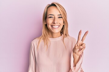 Beautiful blonde woman wearing casual winter sweater smiling with happy face winking at the camera doing victory sign. number two.