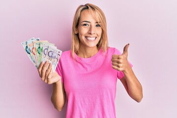 Beautiful blonde woman holding singapore dollars banknotes smiling happy and positive, thumb up doing excellent and approval sign