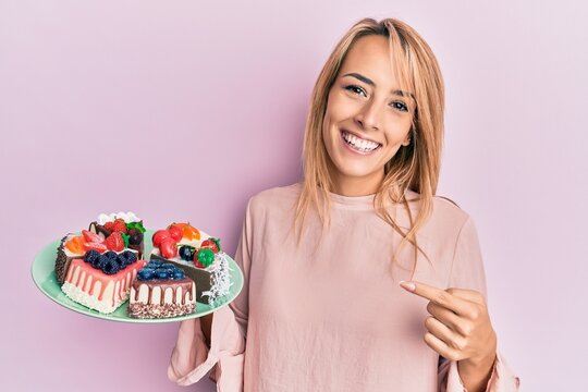 Beautiful blonde woman holding cake slices smiling happy pointing with hand and finger