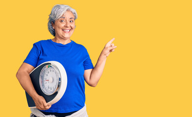 Senior woman with gray hair holding weight machine to balance weight loss smiling happy pointing...