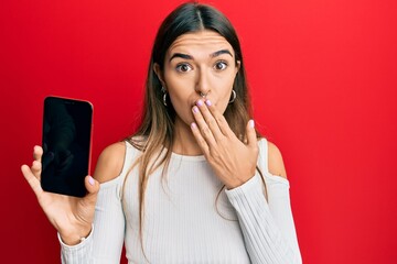 Young hispanic woman holding smartphone showing blank screen covering mouth with hand, shocked and...