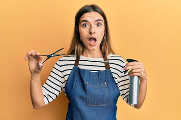 Young hispanic woman wearing hairdresser apron and holding scissors afraid and shocked with...