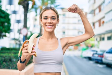 Young cauciasian fitness woman wearing sport clothes training outdoors eating healthy banana and showing proud arm muscle