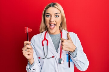 Young caucasian woman wearing doctor uniform holding medical reflex hammer smiling happy and positive, thumb up doing excellent and approval sign