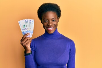 Young african american girl wearing doctor uniform holding 50 colombian pesos looking positive and happy standing and smiling with a confident smile showing teeth