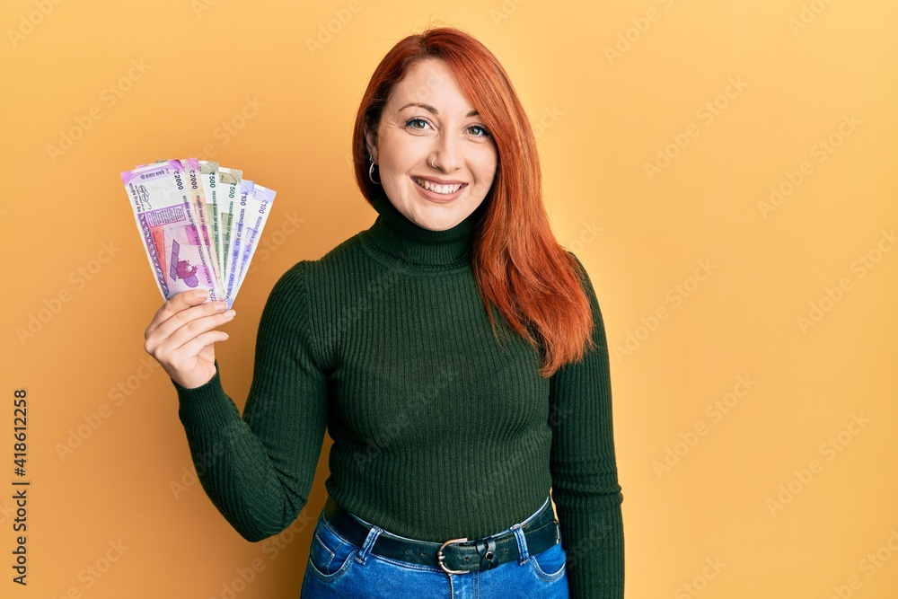 Poster beautiful redhead woman holding indian rupee banknotes looking positive and happy standing and smili