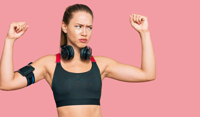 Beautiful blonde woman wearing gym clothes and using headphones showing arms muscles smiling proud. fitness concept.